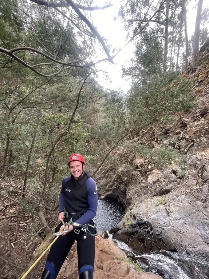 Mann im Neoprenanzug seilt sich über Felsen in ein Wasserbecken ab, umgeben von Bäumen