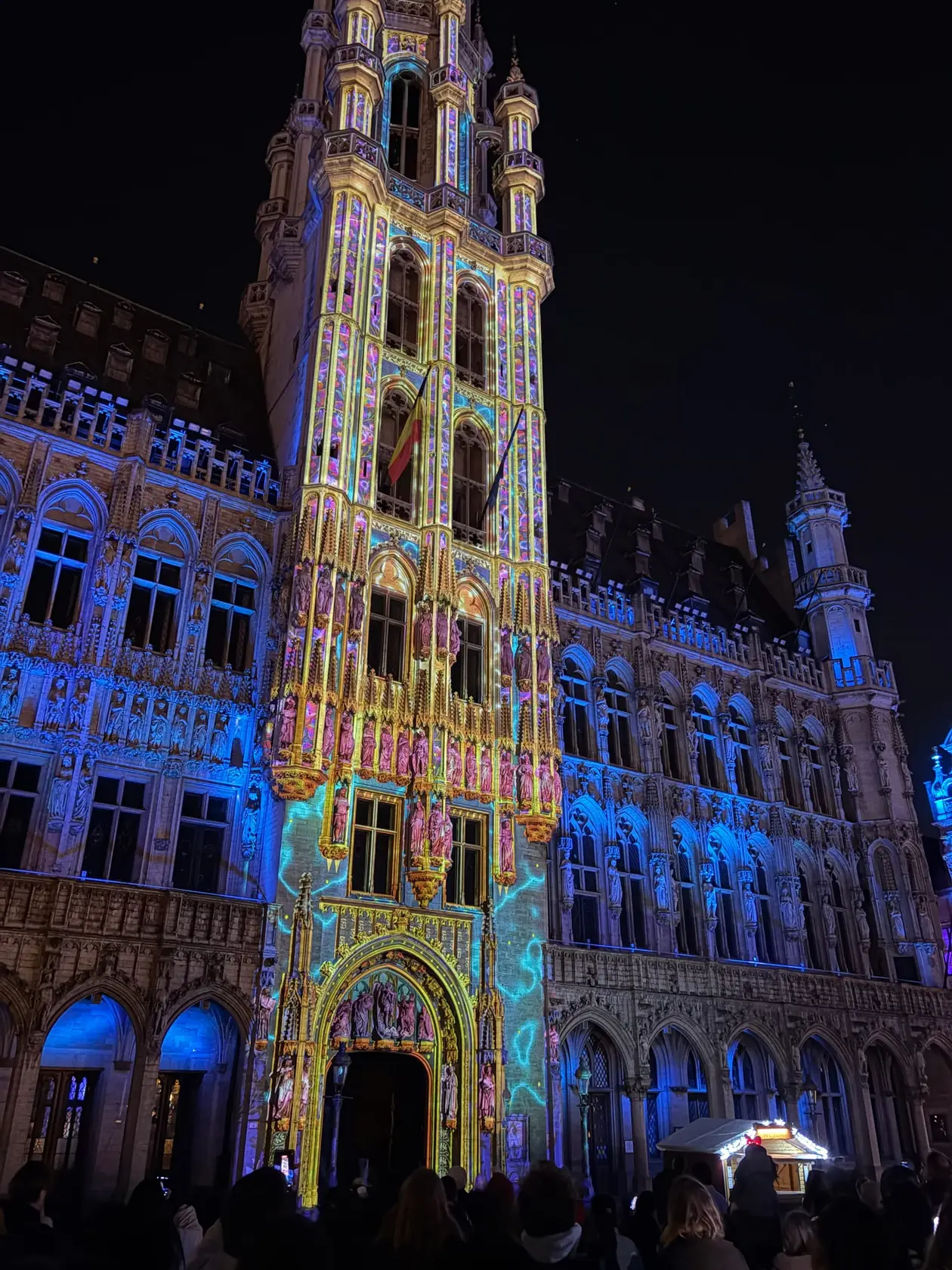 Der Turm des Rathauses am Grand-Place bei Nacht, von der Lichtshow gelb, rosa und blau angestrahlt, unten die Köpfe der Zuschauer