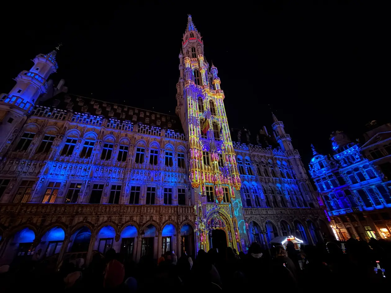 Das gotische Rathaus am Grand-Place bei Nacht, von einer Lichtshow in Blau, Gelb und Rosa angestrahlt, davor die Silhouetten vieler Zuschauer