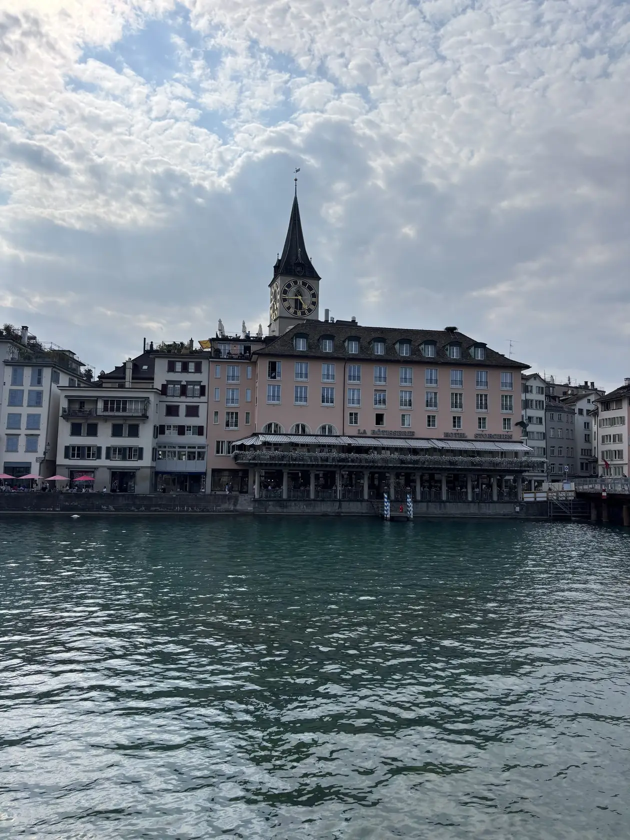 Blick über die Limmat auf eine Häuserzeile mit einem rosa Hotel am Wasser, dahinter der Turm der Kirche St. Peter mit großem Zifferblatt
