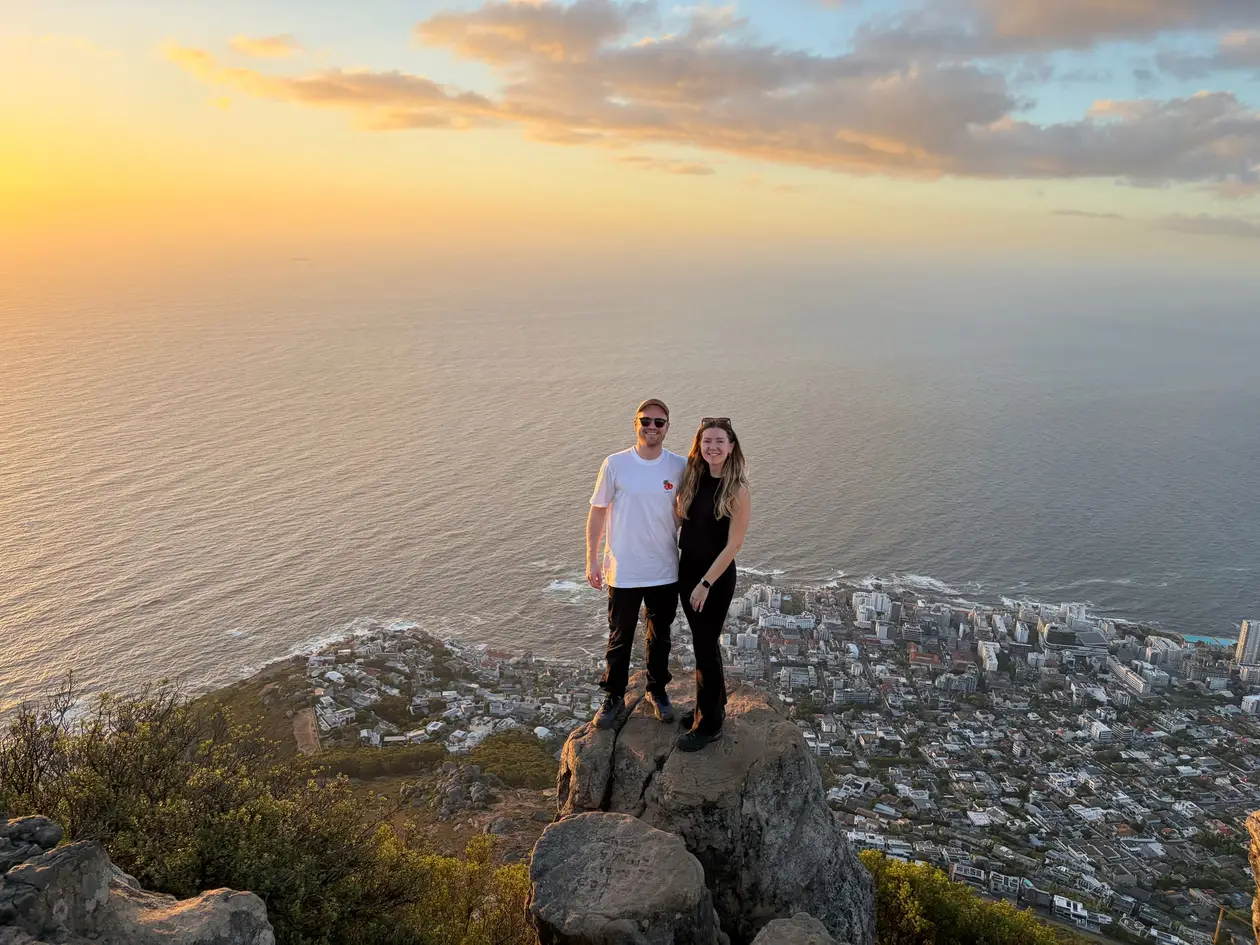 Max und Rebecca stehen bei Sonnenuntergang auf einem Felsen auf dem Lion's Head, dahinter Meer und Küste in warmem Licht