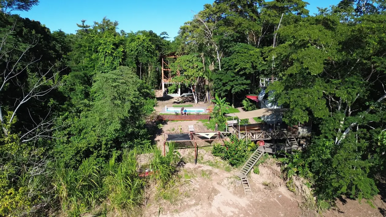 Drohnenblick auf die Lodge, Holzhäuser und ein kleiner Pool zwischen hohen Bäumen, vorne die Uferböschung mit einer Leiter