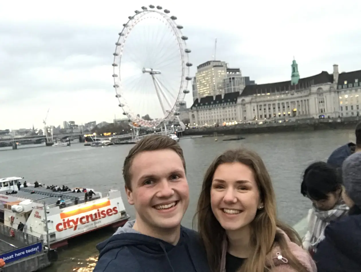 Max und Rebecca lächeln auf der Westminster Bridge in die Kamera, hinter ihnen die Themse, das London Eye und die County Hall