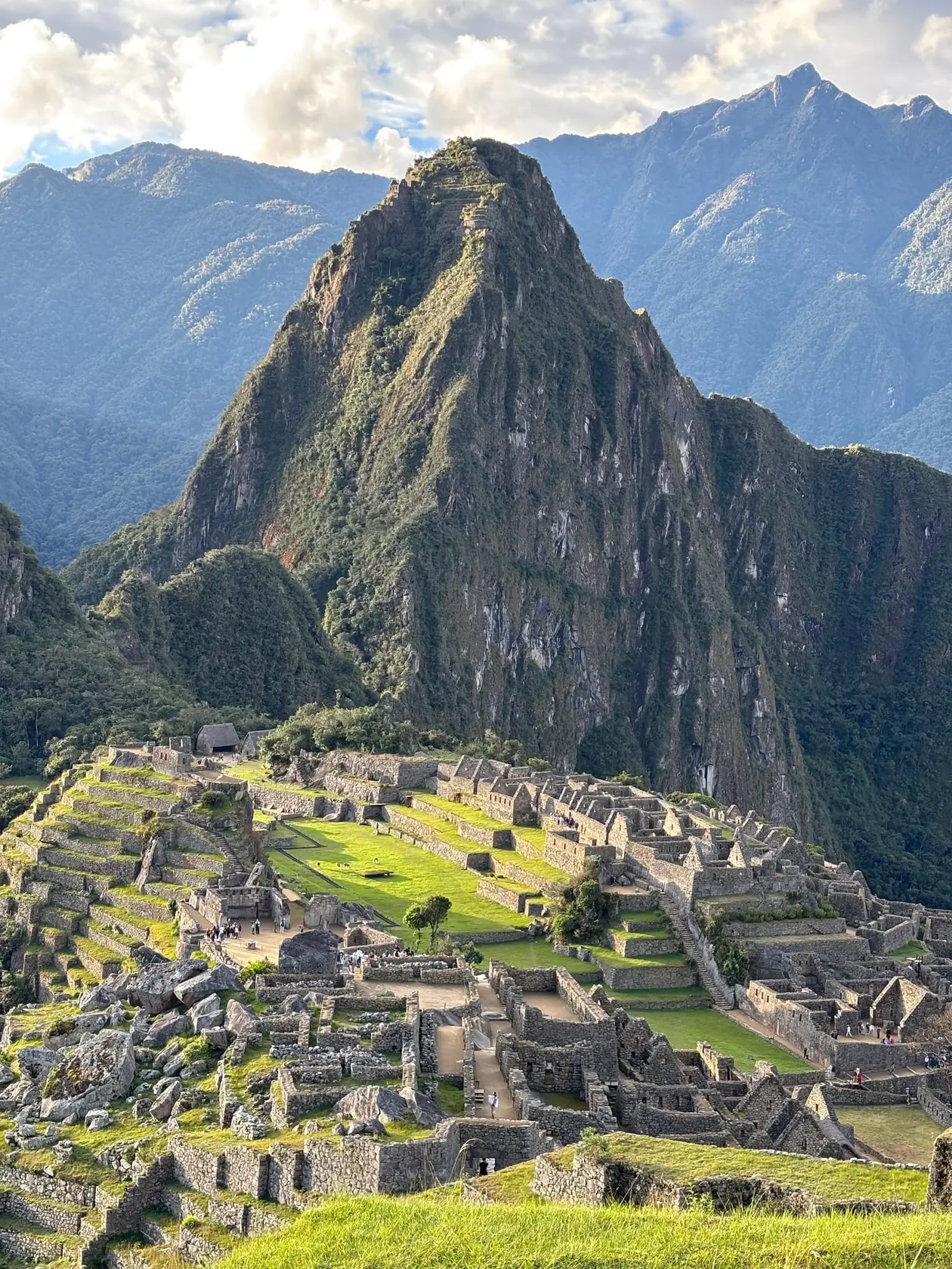 Die Ruinenstadt Machu Picchu mit grünen Plätzen und Terrassen, dahinter der Huayna Picchu im warmen Licht