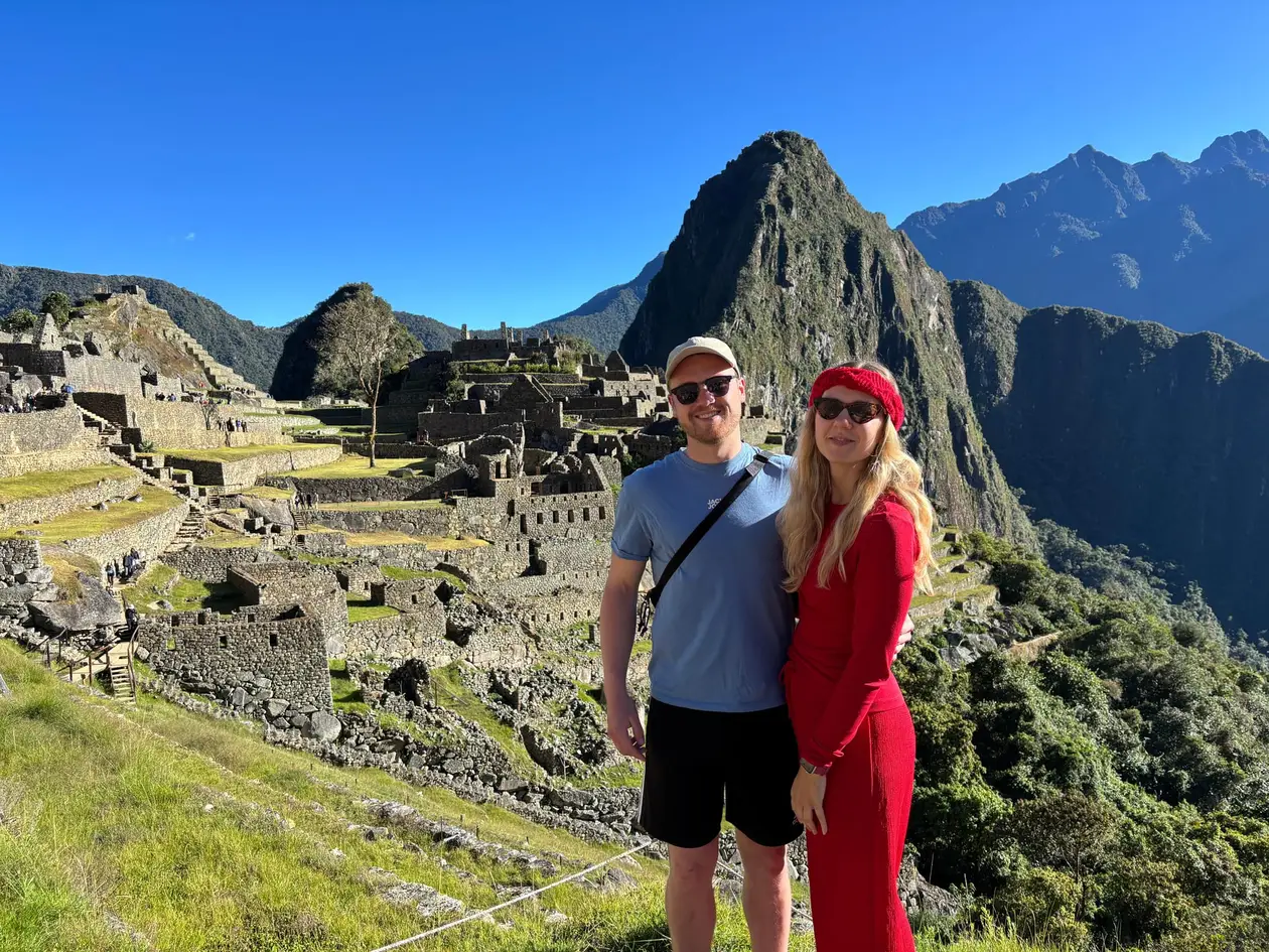 Max und Rebecca stehen auf einer Terrasse oberhalb von Machu Picchu, hinter ihnen die Ruinenstadt und der Huayna Picchu unter wolkenlosem Himmel