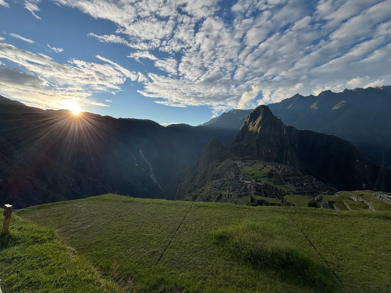 Die Sonne verschwindet hinter einem Bergkamm, im Schatten davor Machu Picchu und der Huayna Picchu, vorne eine grüne Terrasse