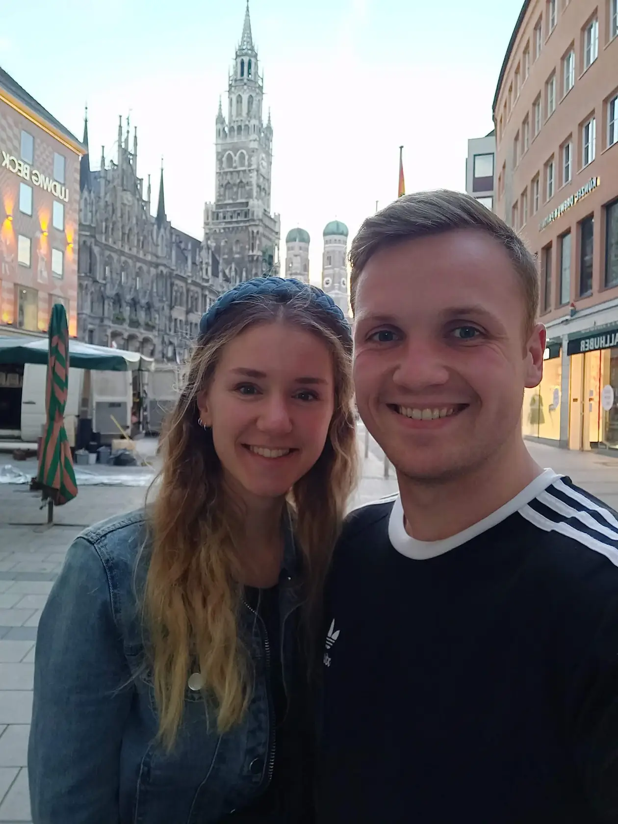 Selfie von Rebecca und Max am Abend auf dem Marienplatz, hinter ihnen das Neue Rathaus mit seinem Turm und die beiden Kuppeltürme der Frauenkirche