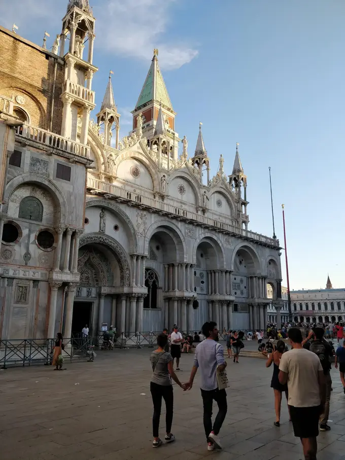 Die Fassade des Markusdoms mit goldenem Abendlicht und der Campanile dahinter, davor ein Paar Hand in Hand auf dem Markusplatz