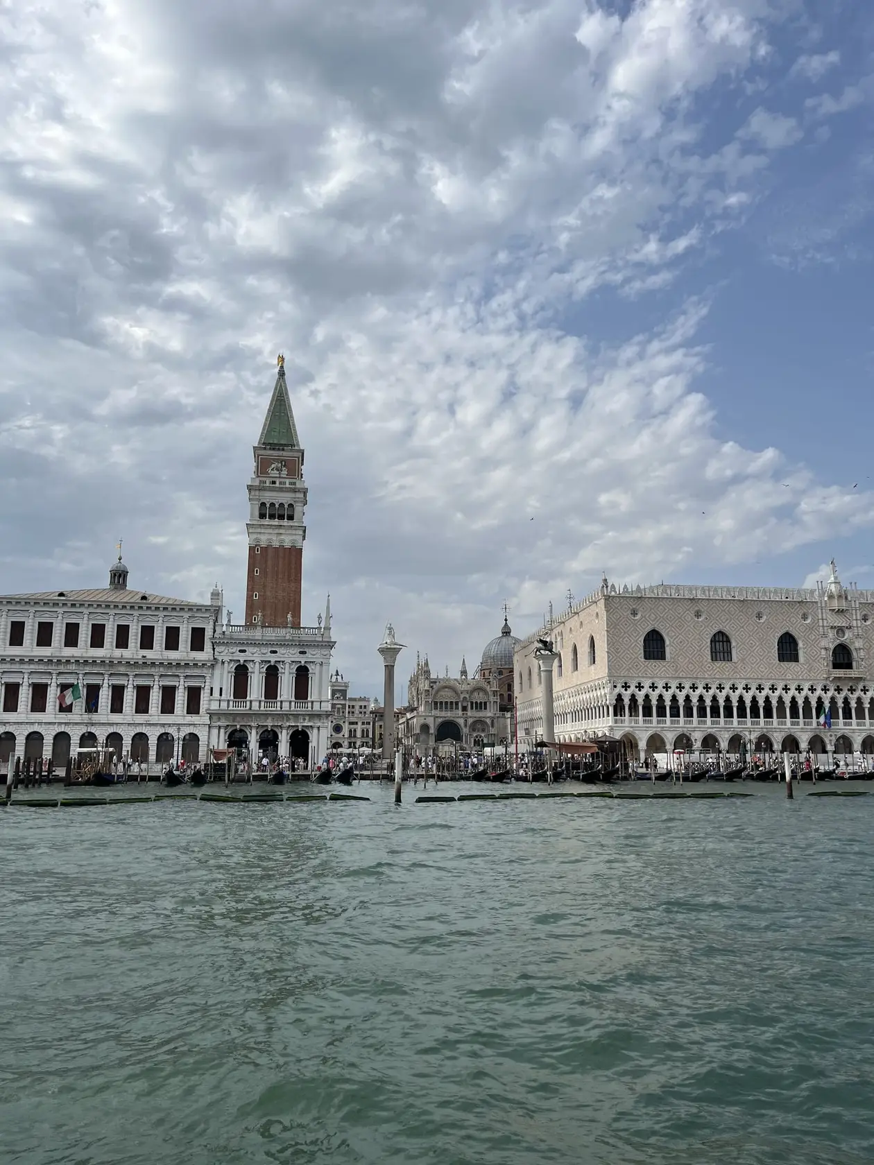 Vom Wasser aus gesehen: die Bibliothek, der Campanile, die Fassade des Markusdoms und der Dogenpalast an der Uferkante, davor Gondeln an Pfählen und grünes Lagunenwasser