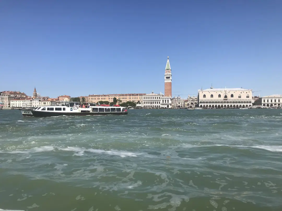 Blick vom Wasser über das Becken auf den Markusplatz mit dem Campanile und dem Dogenpalast, davor ein Ausflugsboot
