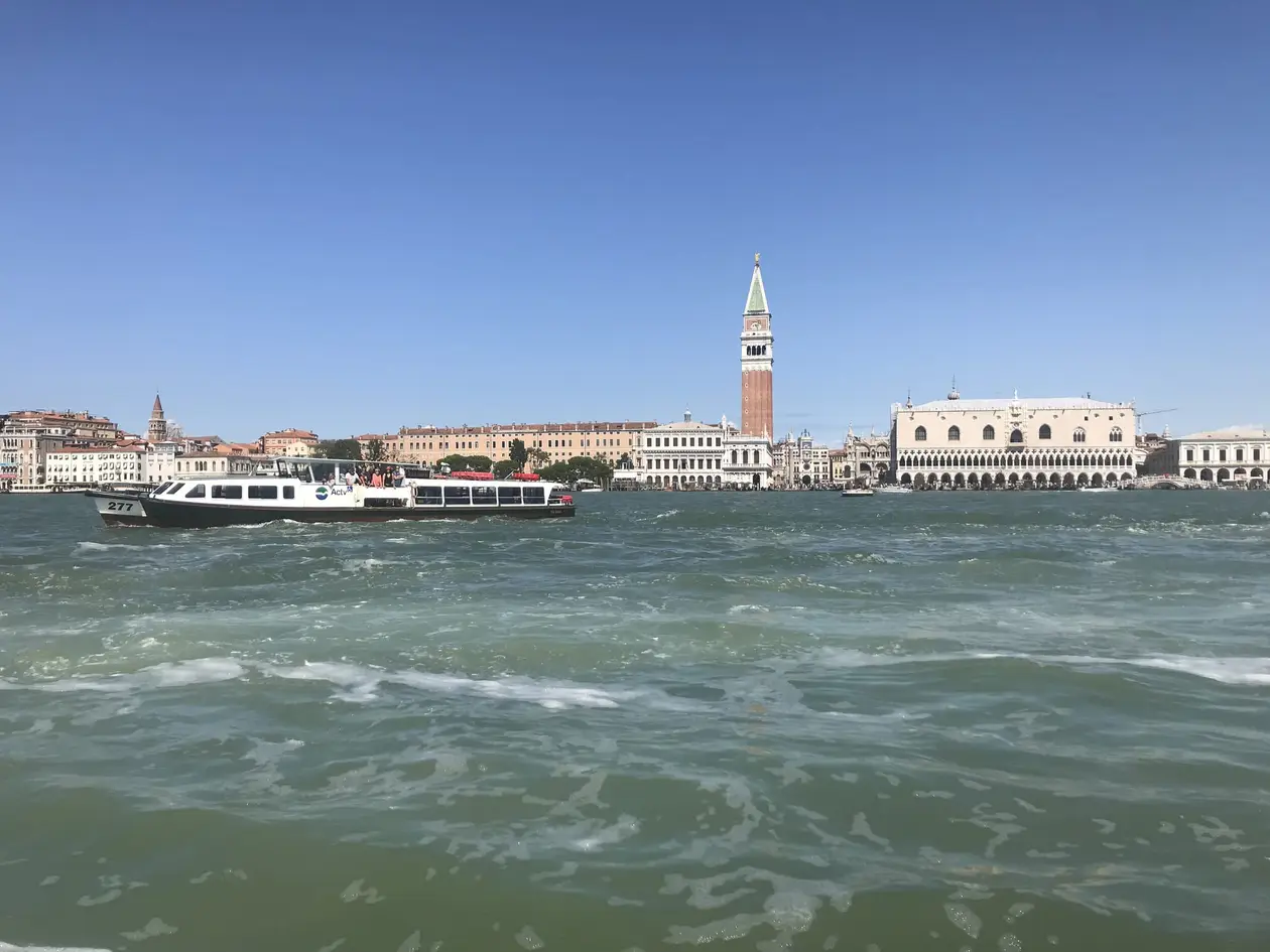 Blick vom Wasser über das Becken auf den Markusplatz mit dem Campanile und dem Dogenpalast, davor ein Ausflugsboot