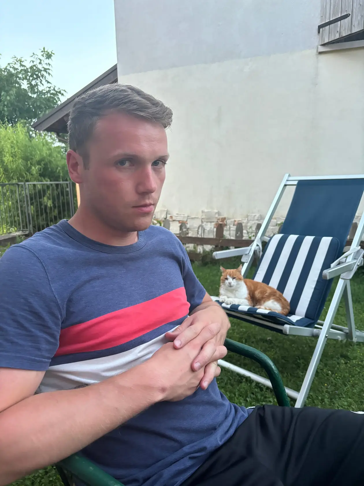 Max sitzt am Abend auf einem Gartenstuhl vor einer hellen Hauswand, hinter ihm liegt eine rot-weiße Katze auf einem gestreiften Liegestuhl
