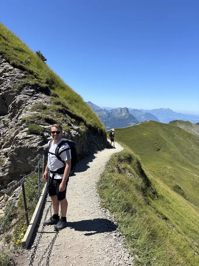 Max steht auf einem schmalen Schotterweg an einer Felswand, rechts führt der Grat weiter, dahinter Berge im Dunst