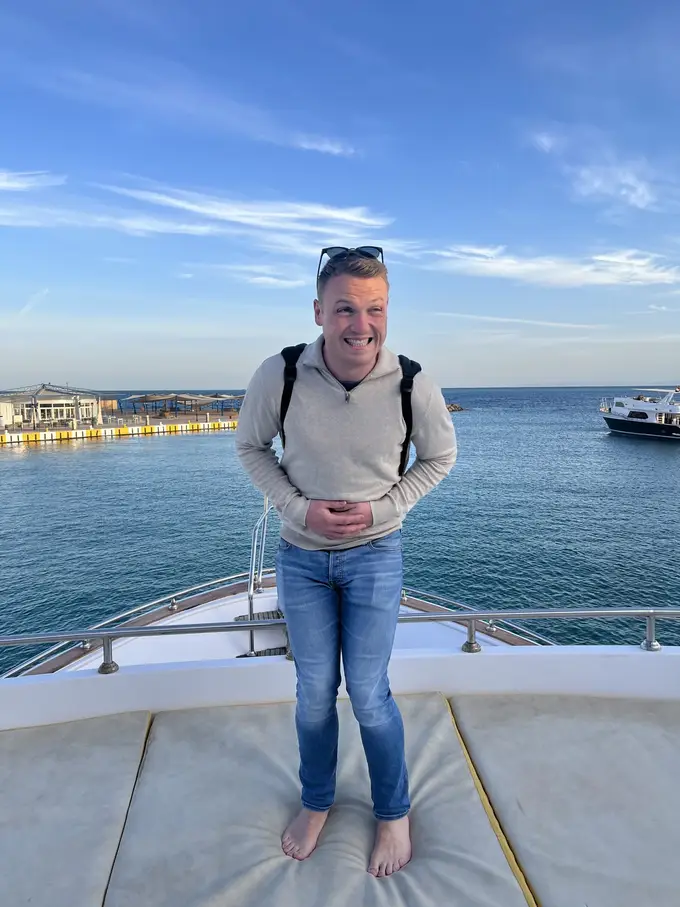 Max steht barfuß im Pullover auf den Polstern am Bug und verzieht das Gesicht, hinter ihm das Hafenbecken und ein weiteres Boot