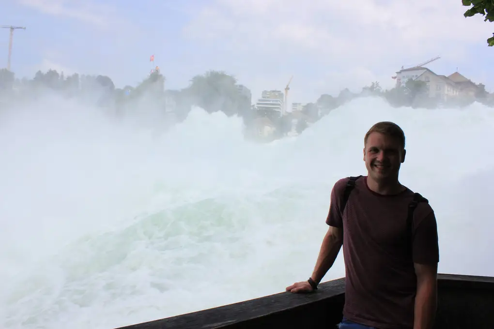 Max lehnt an einem hölzernen Geländer, direkt hinter ihm tobt das Wasser des Rheinfalls in weißer Gischt
