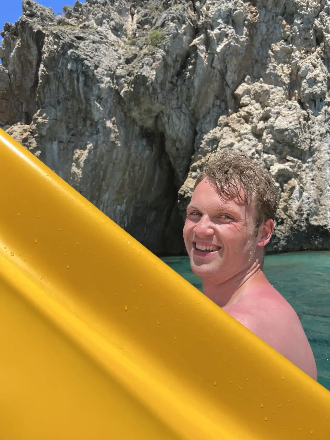 Max lacht aus dem Wasser neben der gelben Rutsche eines Tretboots, dahinter eine Felswand