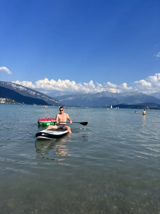 Max sitzt mit Paddel auf dem Board, dahinter der Schwimmring, im Hintergrund Berge unter Quellwolken