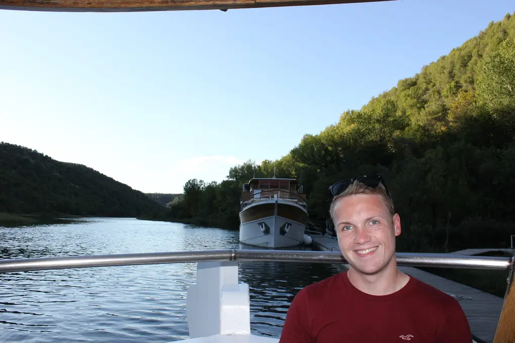 Max im roten T-Shirt an Bord, hinter ihm ein hölzernes Ausflugsboot an einem Steg und das bewaldete Flusstal