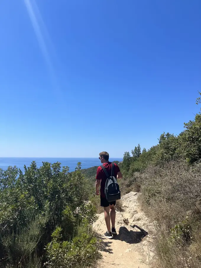Max mit Rucksack auf einem steinigen Pfad zwischen Büschen, vor ihm das Meer