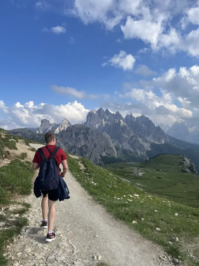 Max von hinten auf einem Schotterweg an einem grünen Hang, vor ihm eine Kette spitzer Felsgipfel unter blauem Himmel mit Wolken