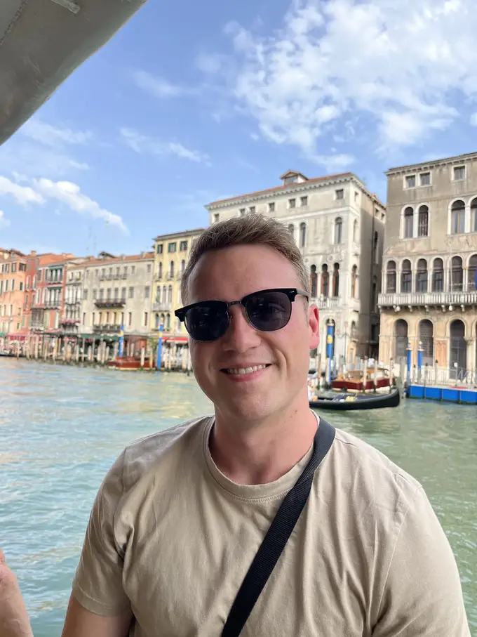 Max mit Sonnenbrille lächelt unter dem Dach eines Vaporetto, hinter ihm der Canal Grande mit Palazzi und einer Gondel