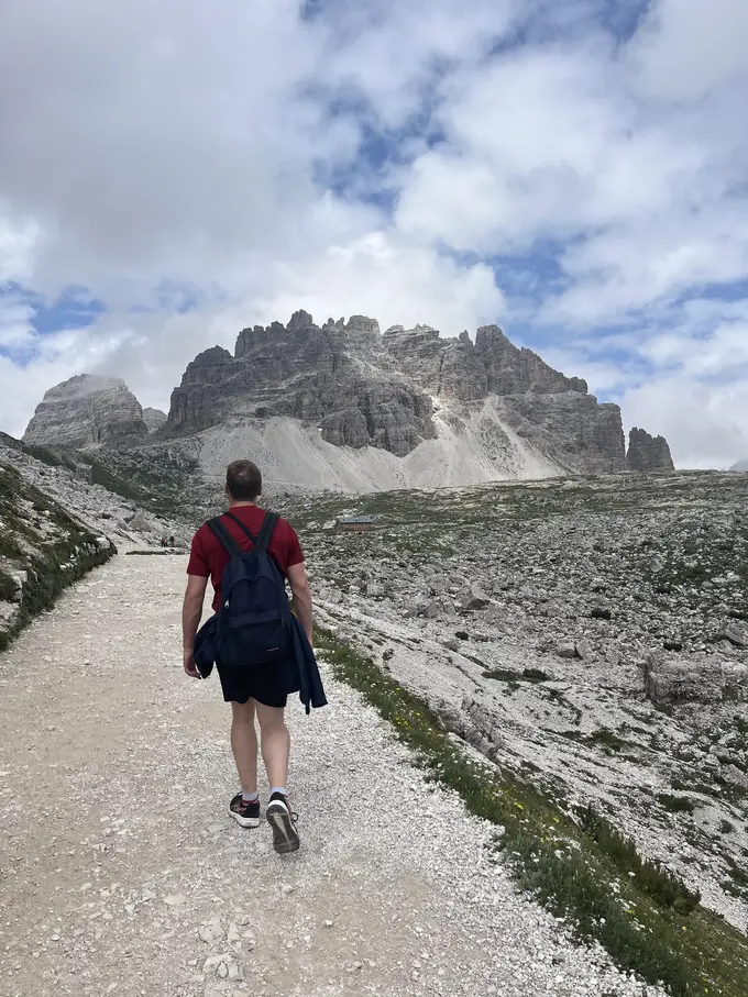 Max von hinten mit Rucksack und Jacke über dem Arm auf einem breiten Schotterweg, vor ihm ein zerklüfteter Felsberg und eine kleine Hütte im Geröll