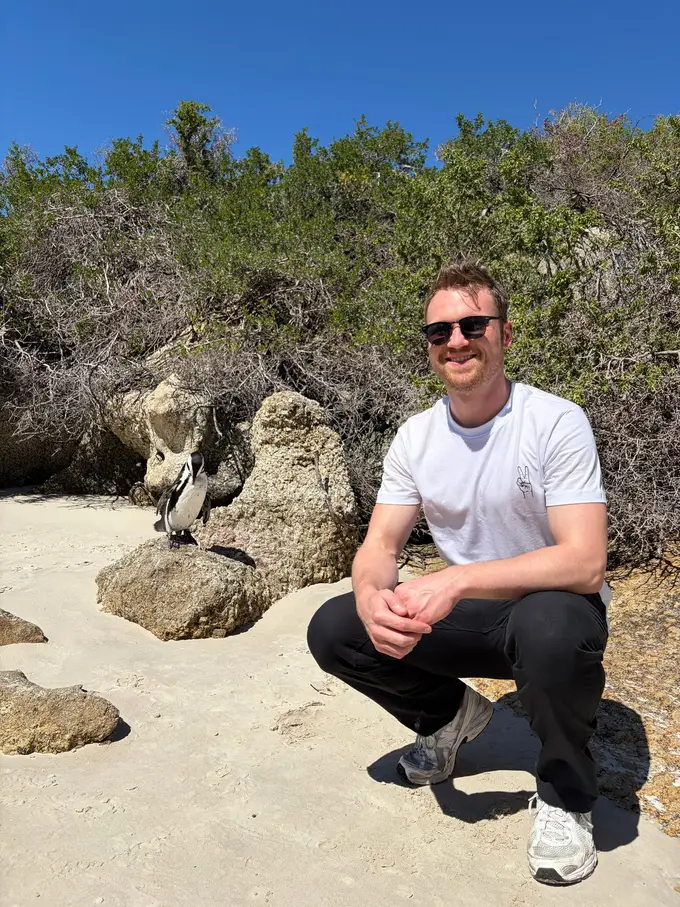 Max hockt lächelnd auf dem Sand, im Hintergrund ein Pinguin zwischen Felsen