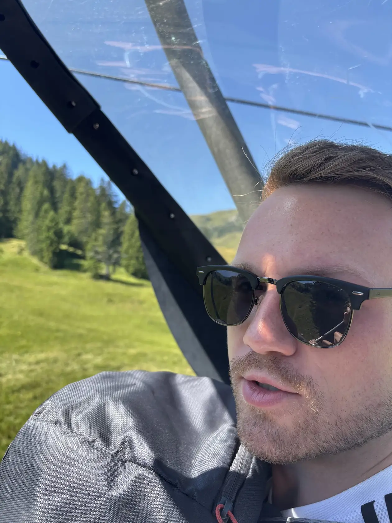 Max sitzt im Sessellift unter einer Glashaube, draußen Nadelbäume und eine Wiese