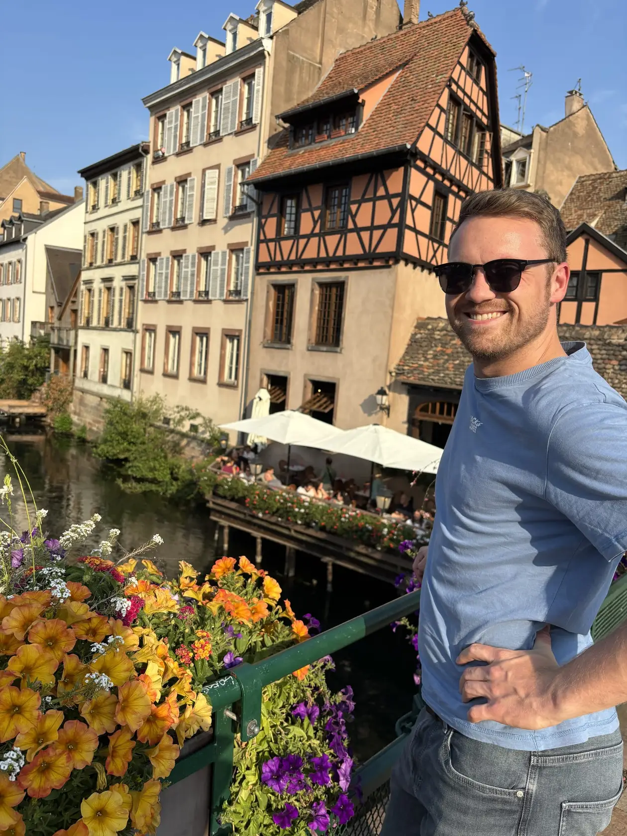 Max steht lächelnd an einem Brückengeländer mit Blumenkästen, hinter ihm ein Kanal und ein orangefarbenes Fachwerkhaus