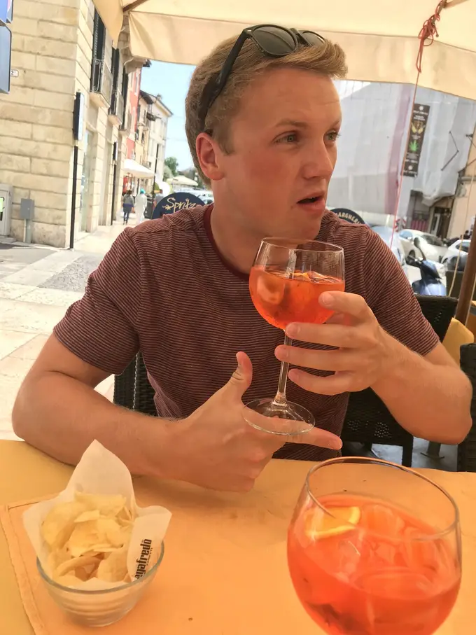 Max sitzt unter einem Sonnenschirm mit einem Aperol in der Hand, auf dem Tisch ein zweites Glas und Chips