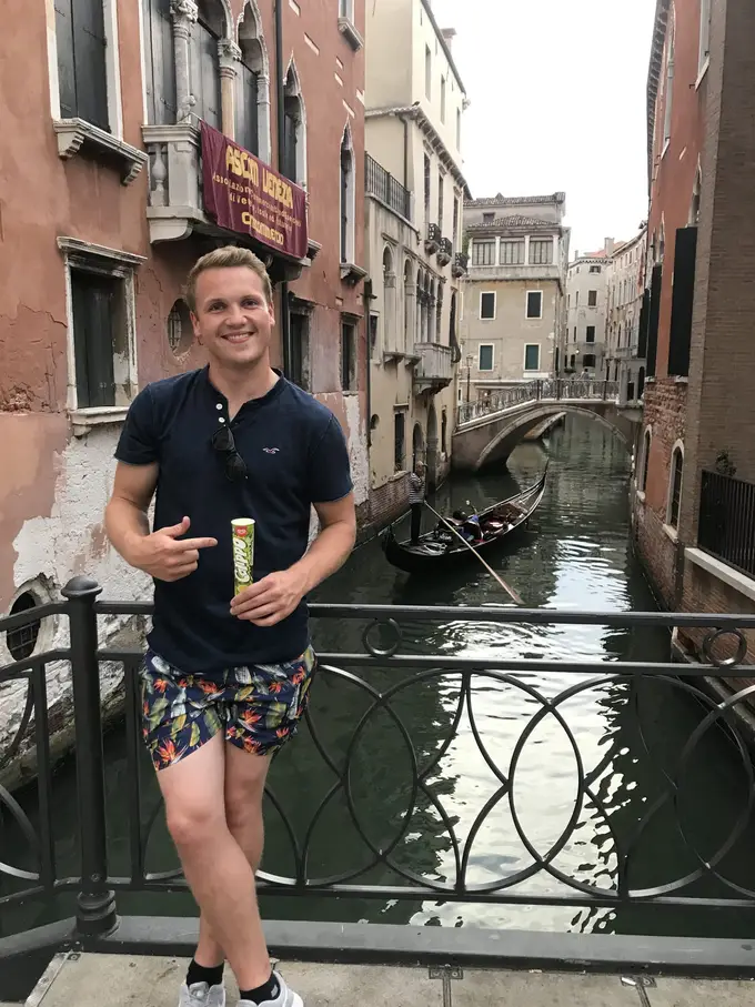 Max steht mit einem Calippo auf einer Brücke über einem schmalen Kanal, hinter ihm eine Gondel