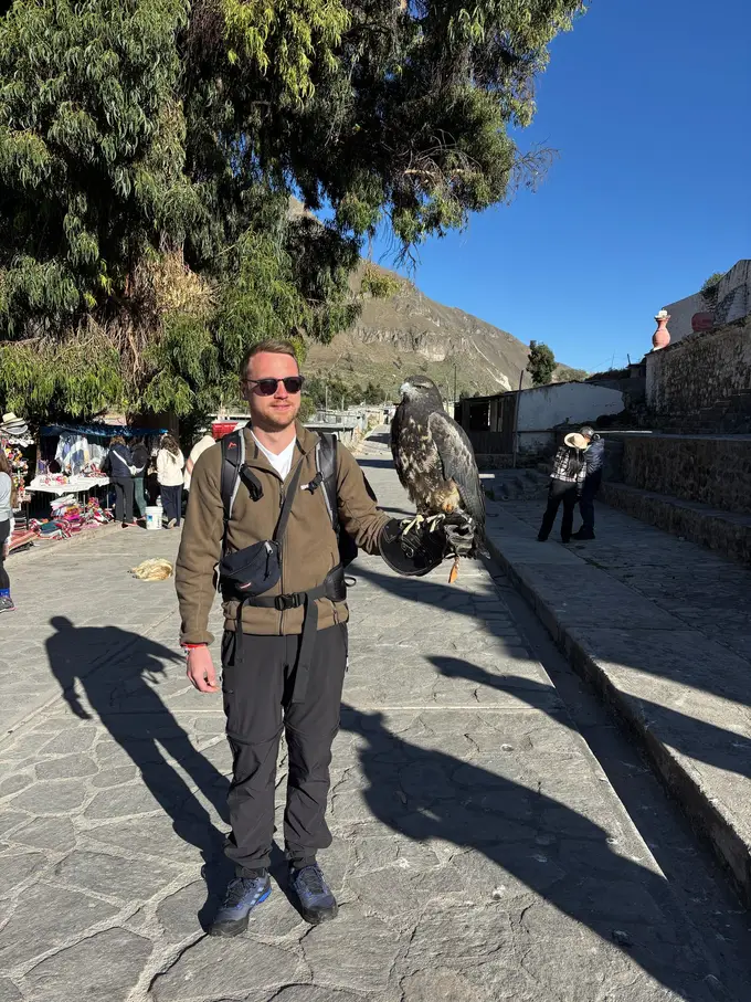 Max steht auf dem Dorfplatz von Maca und hält einen großen Greifvogel auf dem behandschuhten Arm