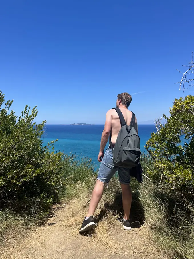 Max mit nacktem Oberkörper und Rucksack auf einem Pfad zwischen Büschen, vor ihm das Meer