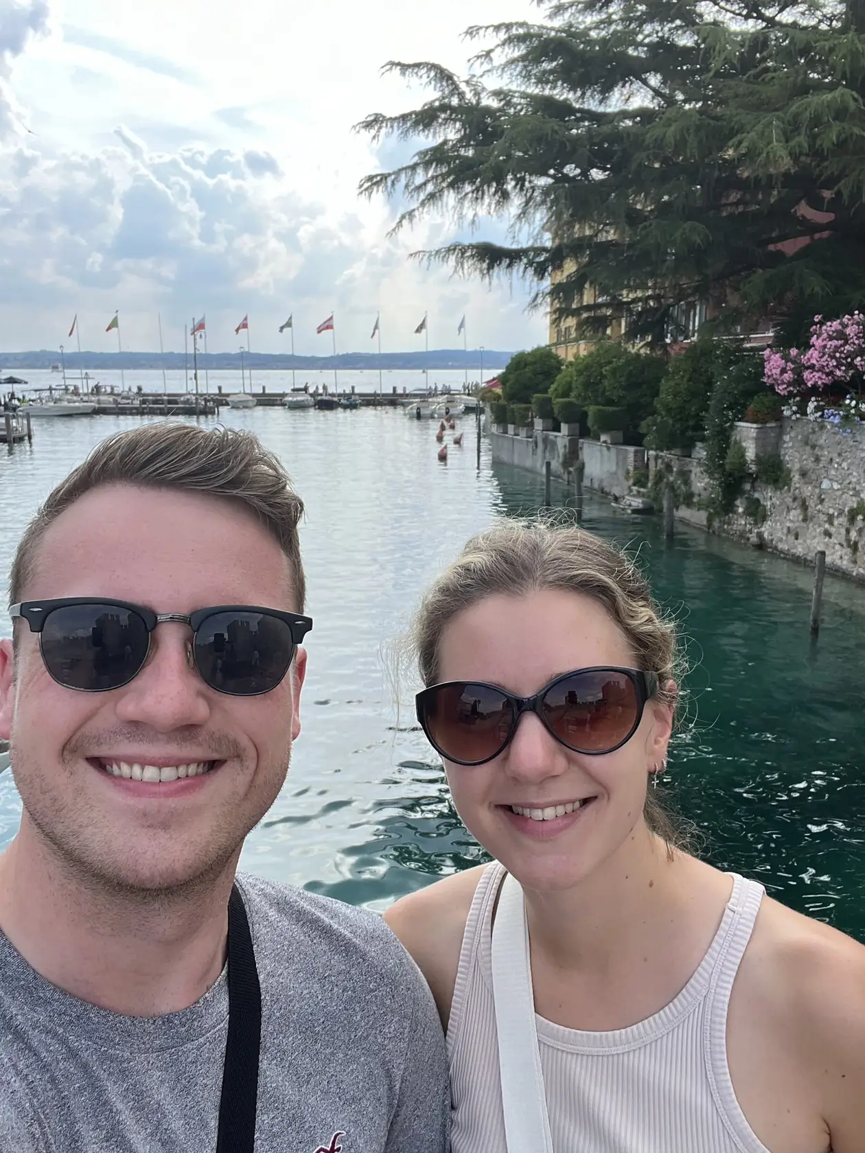 Selfie von Max und Rebecca mit Sonnenbrillen vor einem Hafenbecken mit grünem Wasser, dahinter Boote, eine Reihe Fahnen und eine Zeder