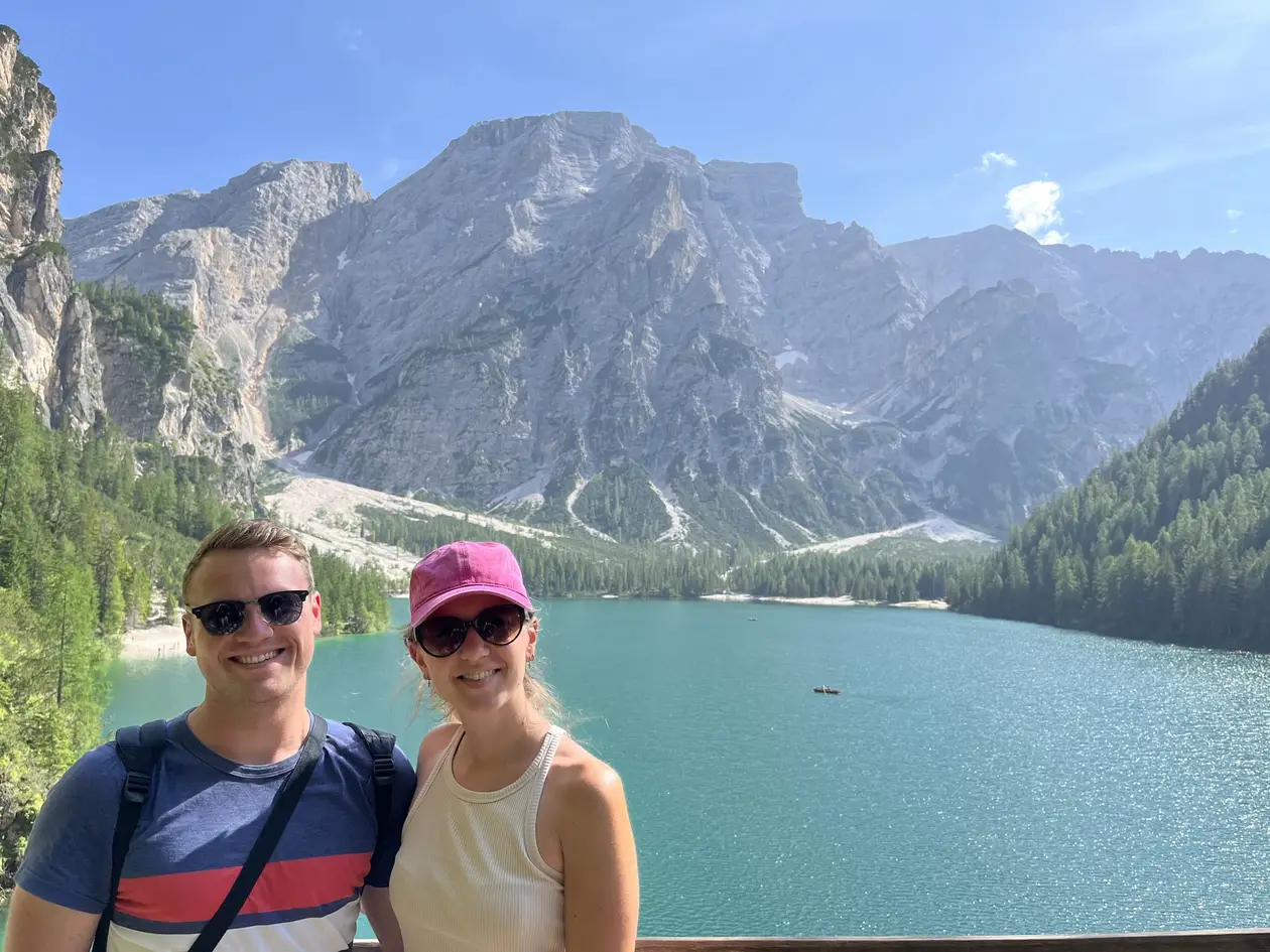 Max und Rebecca lächeln vor dem türkisgrünen Pragser Wildsee, dahinter ein großes helles Felsmassiv und bewaldete Hänge, auf dem Wasser ein einzelnes Ruderboot
