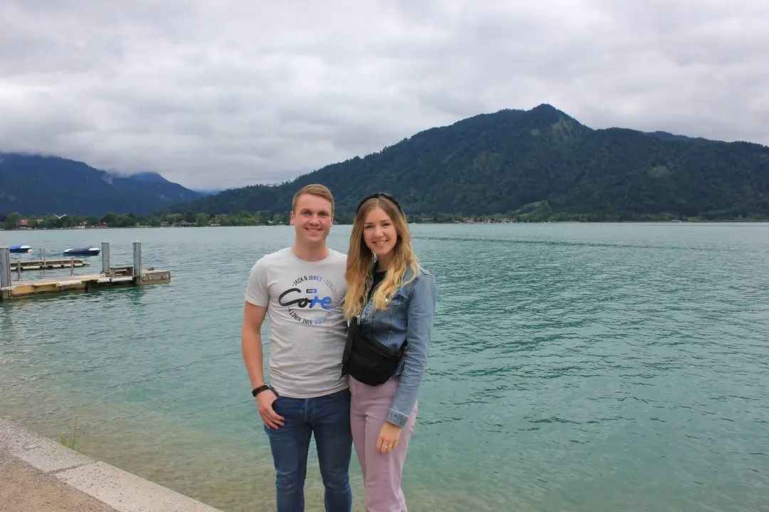 Max und Rebecca stehen am Ufer vor dem türkisgrünen See, links ein Steg, dahinter bewaldete Berge unter Wolken