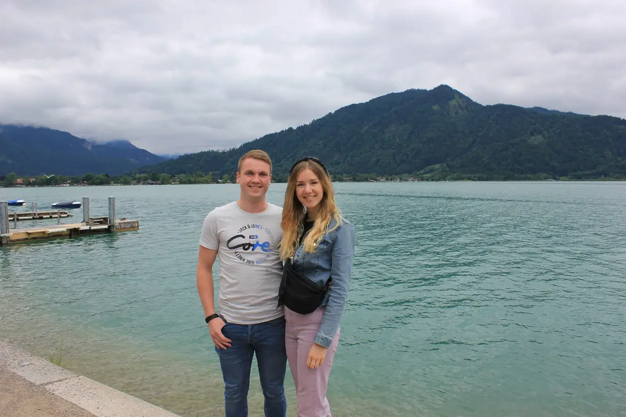 Max und Rebecca stehen am Ufer vor dem türkisgrünen See, links ein Steg, dahinter bewaldete Berge unter Wolken