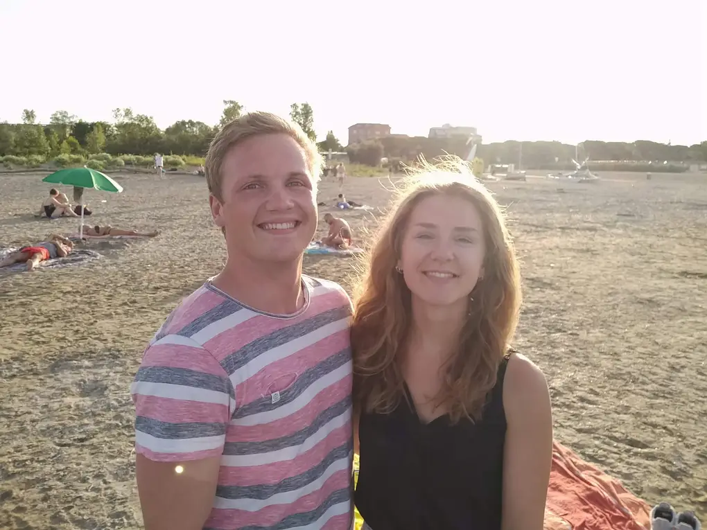 Max und Rebecca im Gegenlicht auf dem Sandstrand, im Hintergrund Sonnenschirme und Badegäste