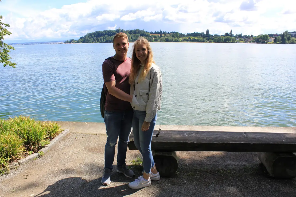 Max und Rebecca stehen neben einer Holzbank am Ufer, hinter ihnen der See und ein bewaldetes Ufer