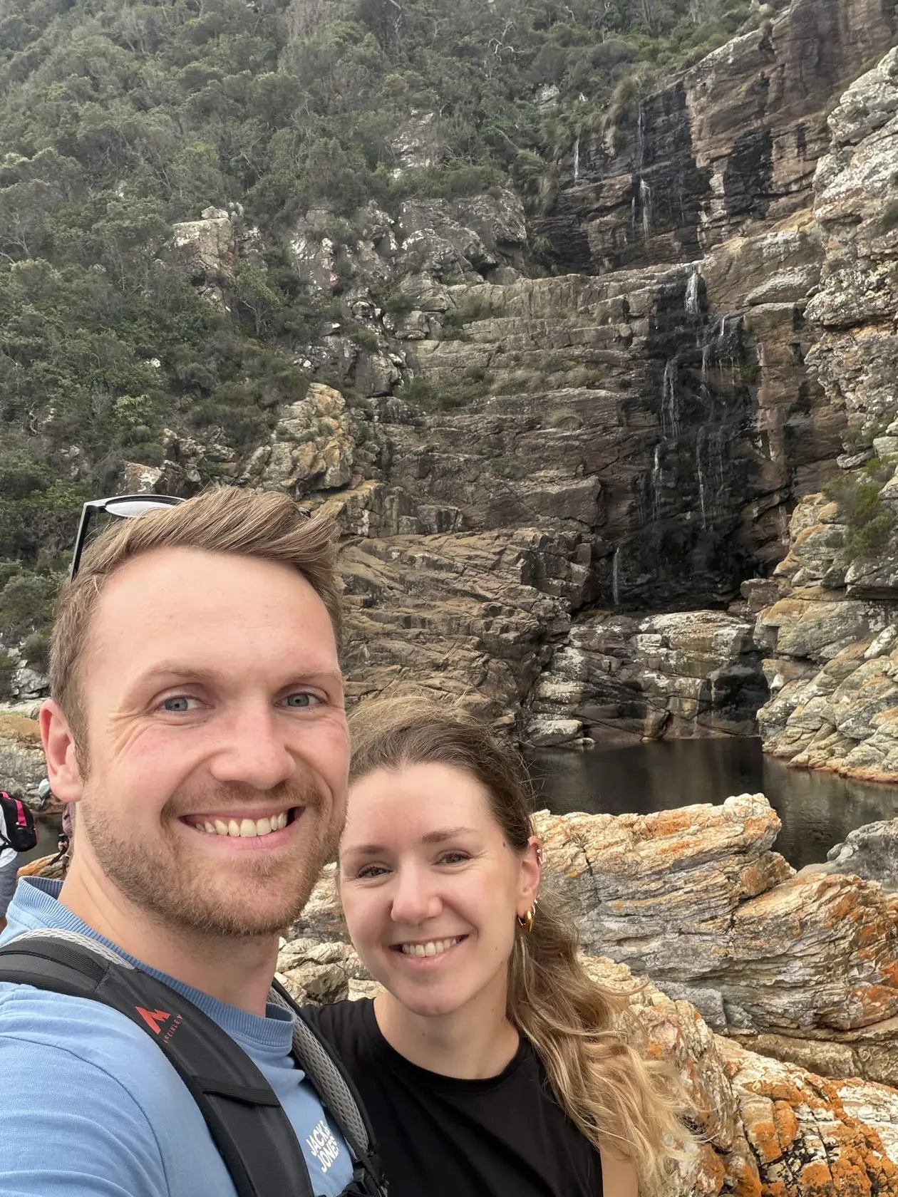Max und Rebecca lächeln vor einem Wasserfall, der in ein dunkles Becken zwischen Felsen fällt