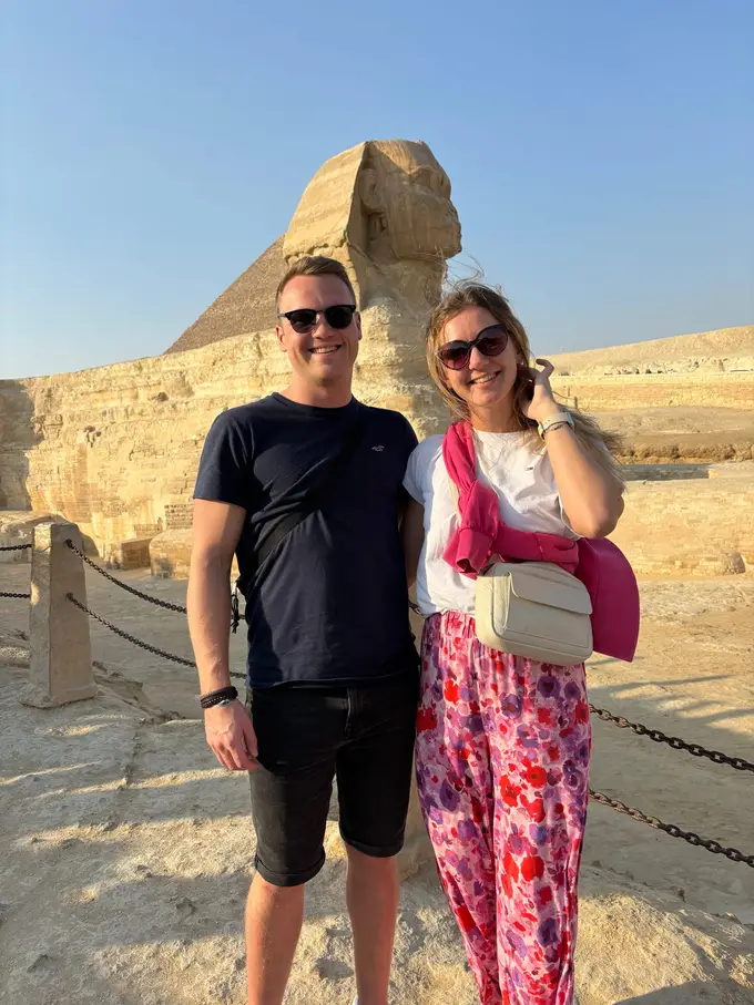 Max und Rebecca stehen lächelnd vor einer Absperrkette, direkt hinter ihnen die Sphinx und dahinter eine Pyramide