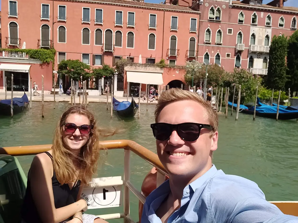 Selfie von Max und Rebecca an der Reling eines Boots, hinter ihnen Gondeln vor einer roten Palastfassade