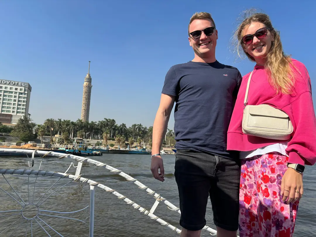 Max und Rebecca stehen lächelnd am Bug eines Boots auf dem Nil, hinter ihnen das Ufer mit Palmen, ein Hotelhochhaus und der Cairo Tower