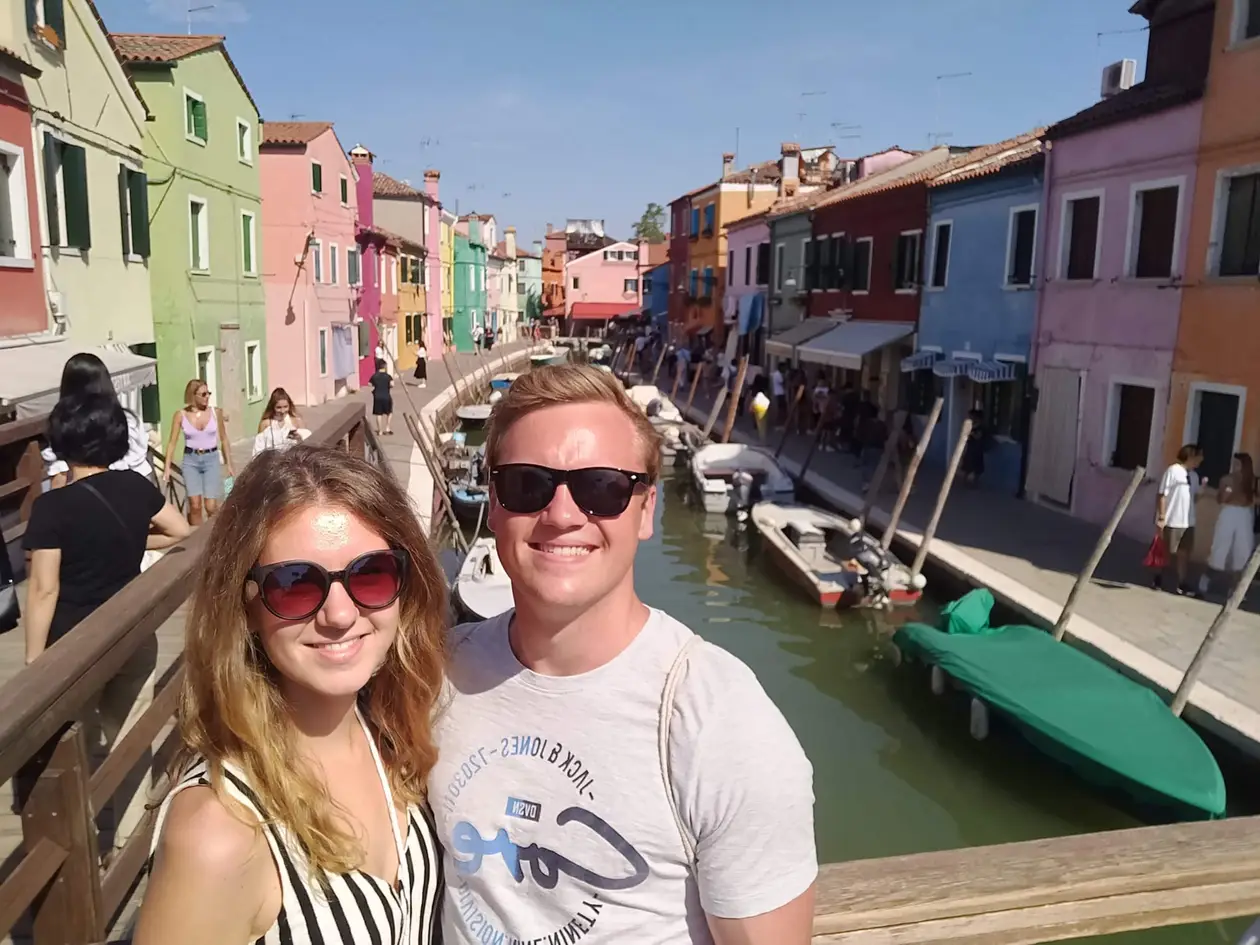 Selfie von Rebecca und Max mit Sonnenbrillen auf einer Holzbrücke, hinter ihnen ein Kanal mit Booten und Häuser in Grün, Rosa, Rot und Blau