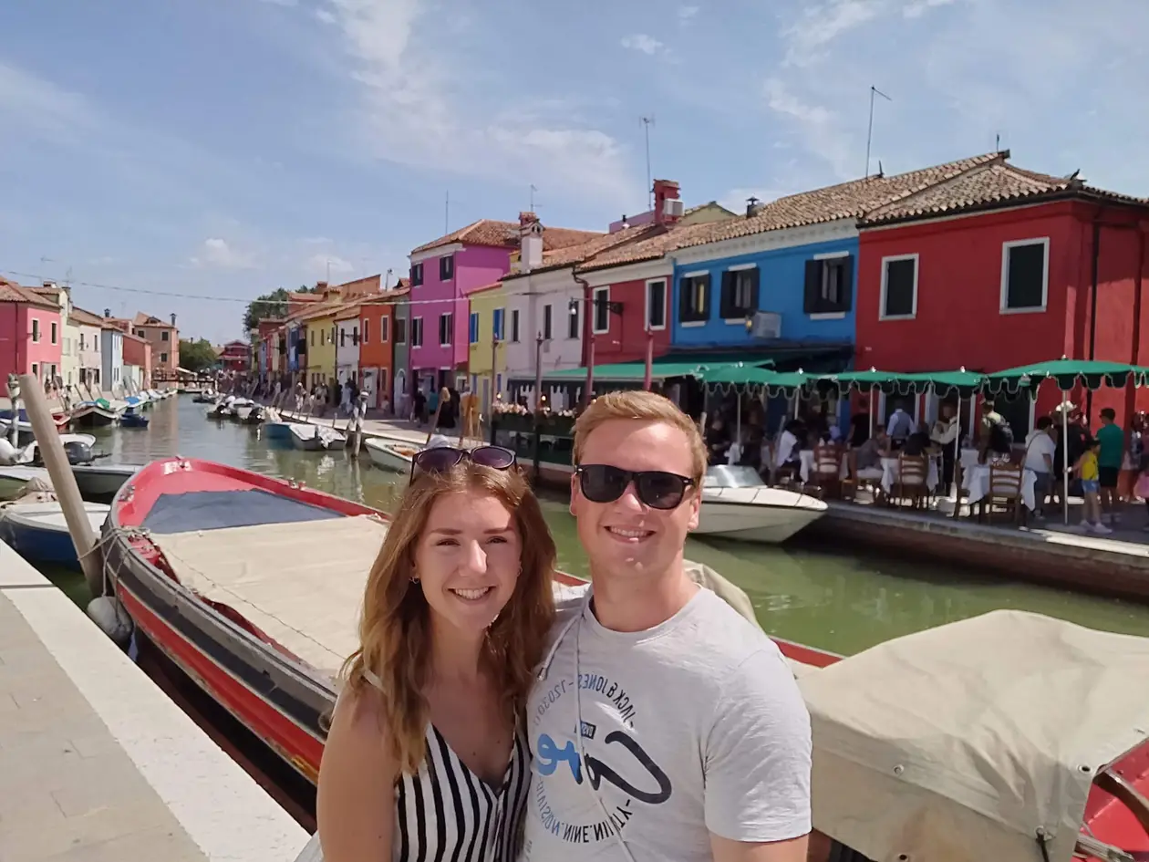 Rebecca und Max mit Sonnenbrillen an einem Kanal auf Burano, hinter ihnen Boote und eine Reihe roter, blauer und rosa Häuser