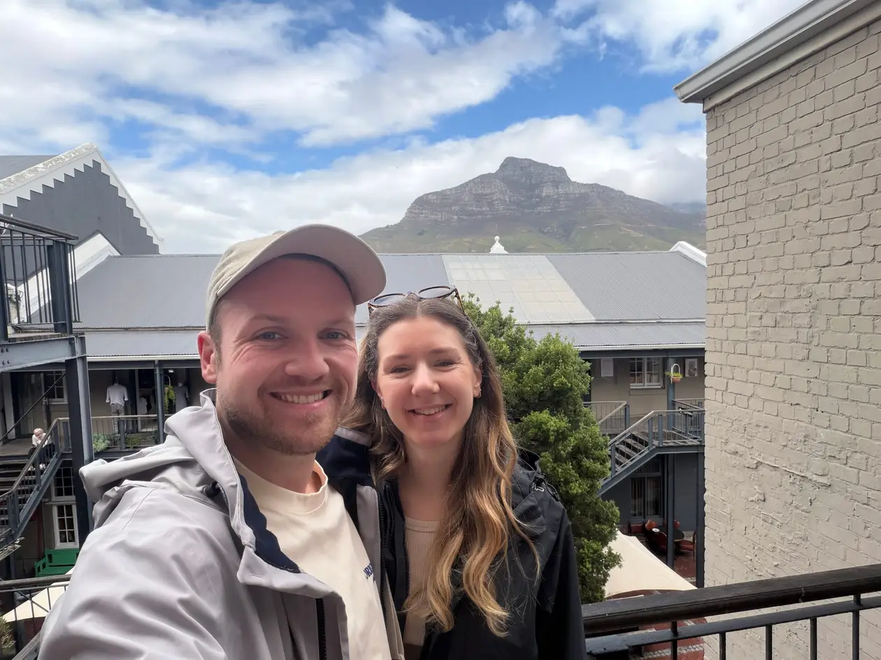 Max und Rebecca lächeln in die Kamera auf einer Empore der Old Biscuit Mill, im Hintergrund der Tafelberg