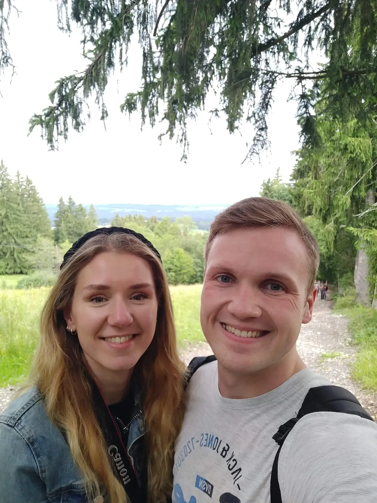 Selfie von Rebecca und Max unter einer Fichte an einem Schotterweg, hinter ihnen Wiesen, Wald und eine weite, dunstige Landschaft