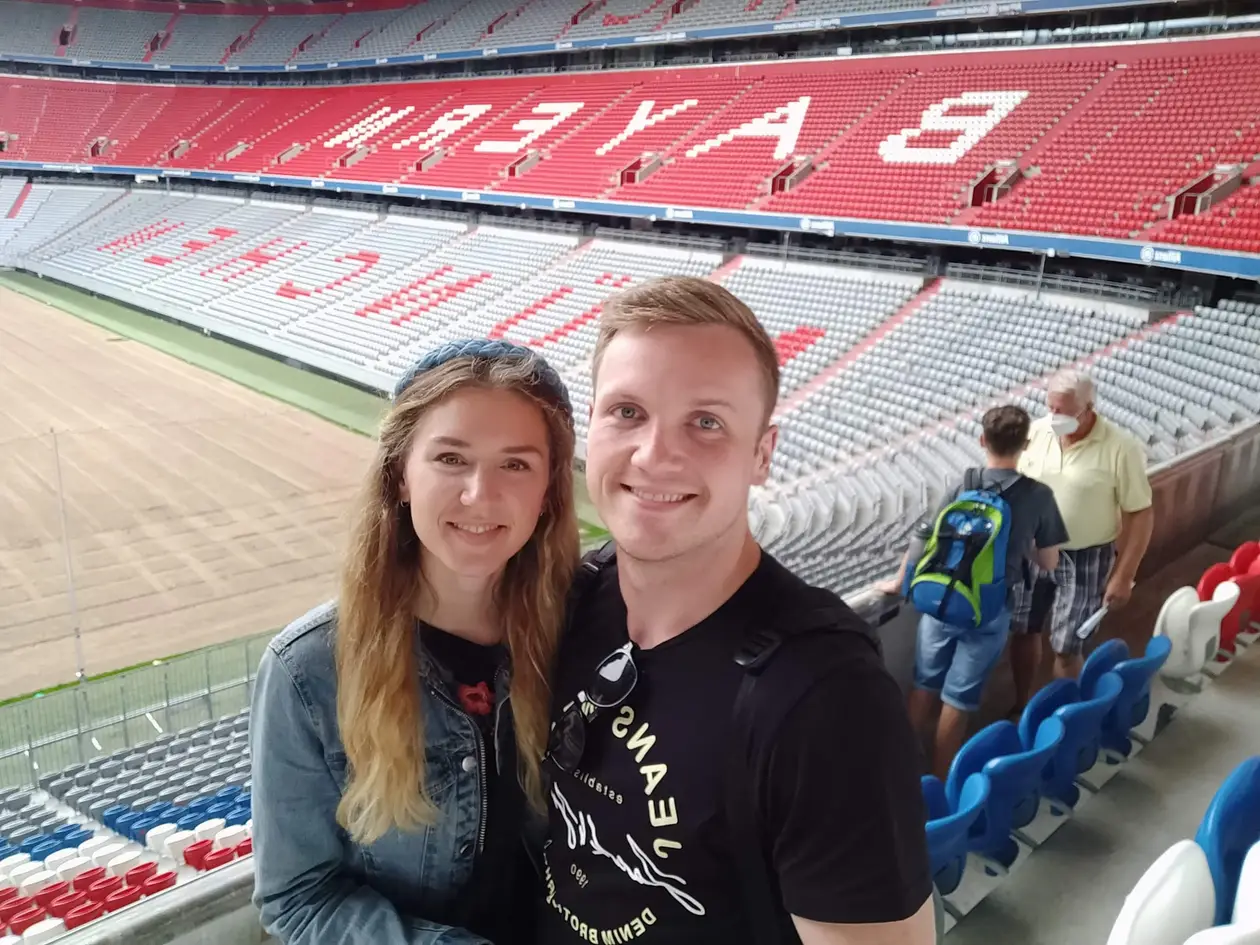 Selfie von Rebecca und Max auf der Tribüne der leeren Arena, dahinter rote und graue Sitzreihen und ein Spielfeld, auf dem der Rasen gerade neu verlegt wird