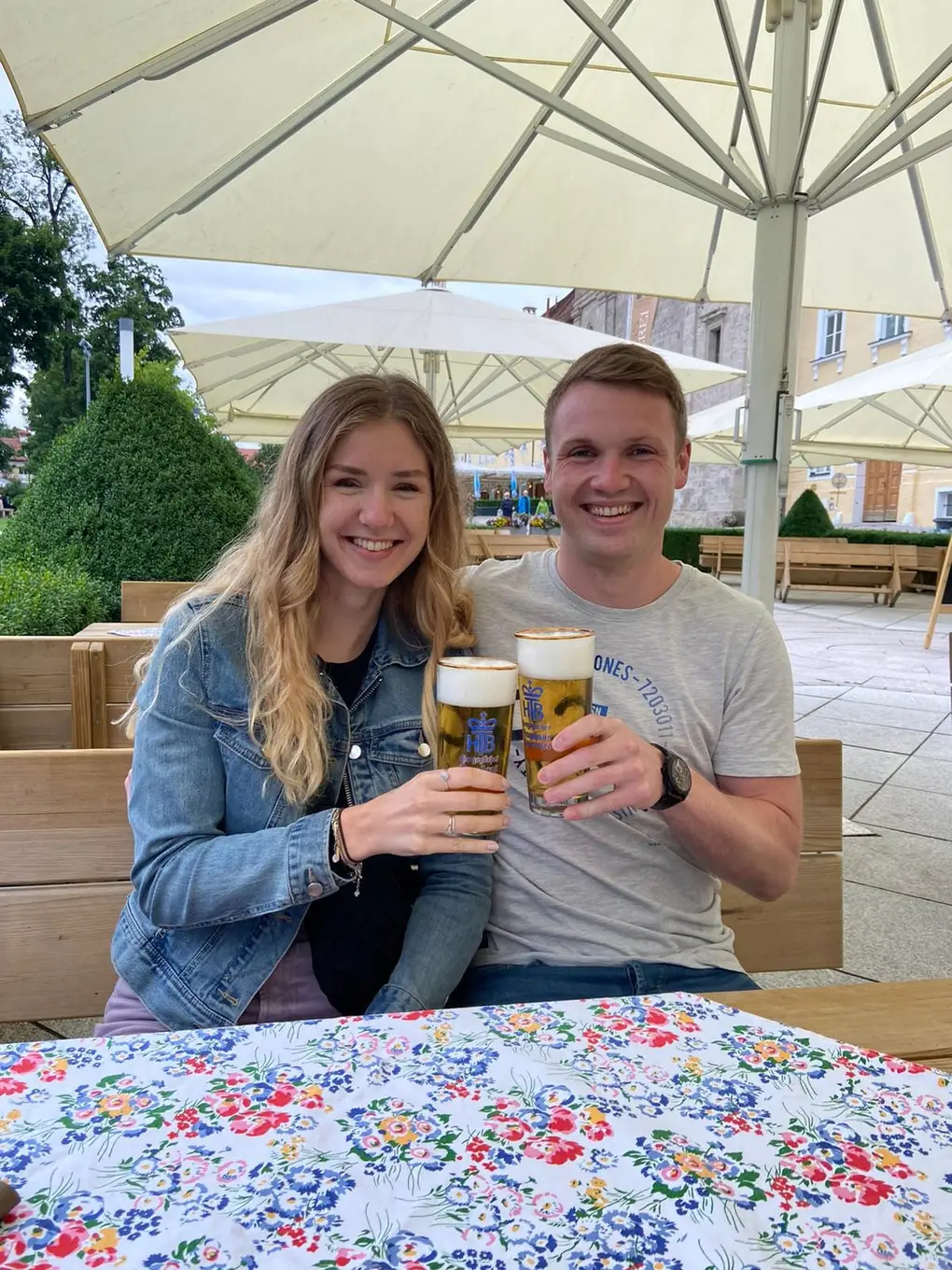 Rebecca und Max stoßen unter weißen Sonnenschirmen mit zwei Gläsern Bier an, vorne eine bunt geblümte Tischdecke