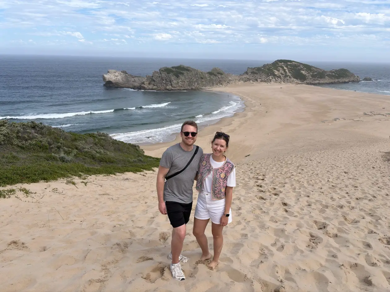 Max und Rebecca stehen auf einer Sanddüne mit Blick auf eine Bucht mit Felsinsel